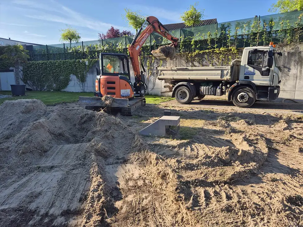 Kleiner Bagger lädt Erde in Muldenkipper auf einer Baustelle mit Sandhaufen und Betonplatte im Hintergrund