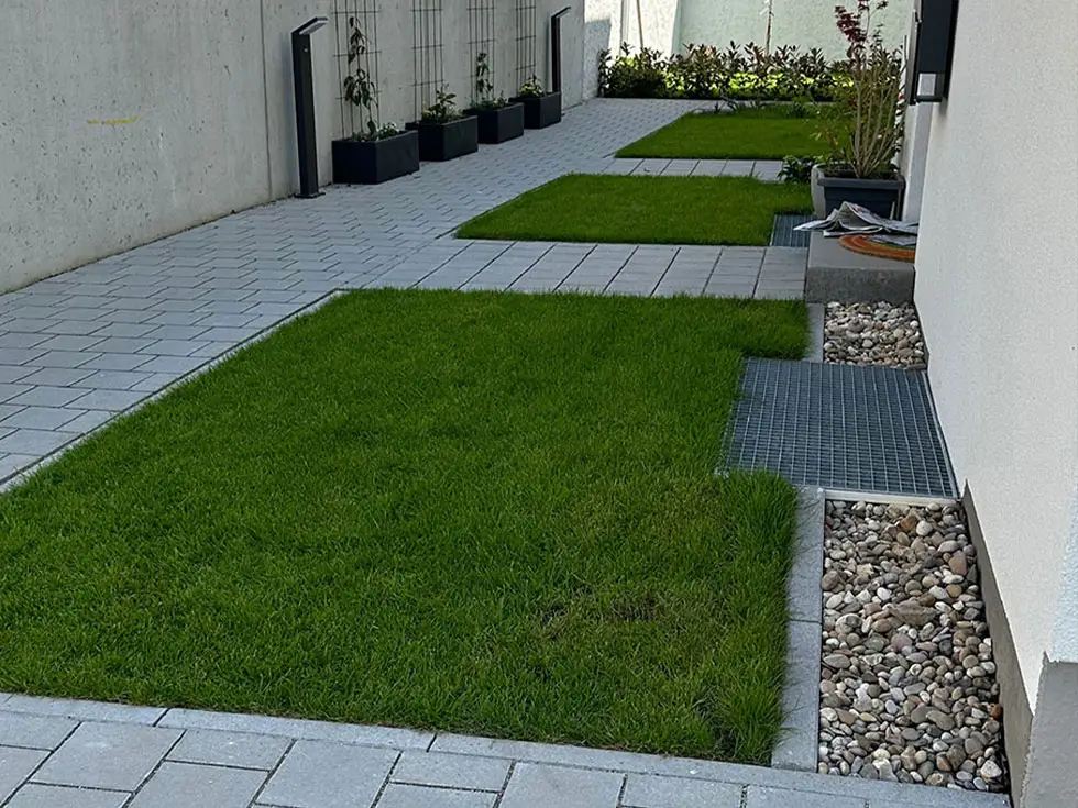 Gepflasterter Weg mit rechteckigen Grünflächen und Pflanzkübeln entlang einer Hauswand