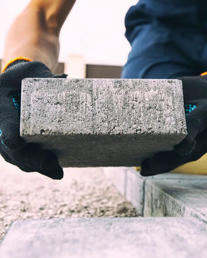 Person mit schwarzen Handschuhen hält einen rechteckigen Pflasterstein hochkant über einem Boden aus Pflastersteinen